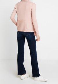Frau trägt dunkelblaue, schmal geschnittene Jeans, ein hellrosa, geripptes Langarmoberteil und weiße Sneakers, stehend vor einem schlichten weißen Hintergrund.