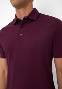 Polo in borgogna con colletto classico, tre bottoni e tessuto texturizzato. Le maniche corte completano il design per una vestibilità rilassata.