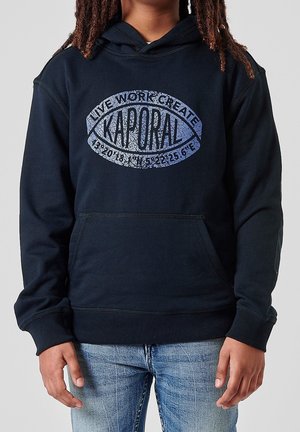 Person trägt einen schwarzen Hoodie mit dem Text "KAPORAL" und Koordinaten in einem fischförmigen Logo, kombiniert mit hellblauen Jeans.