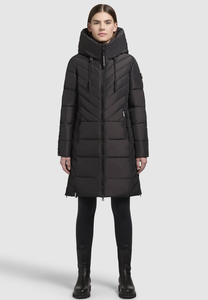 Schwarzer, gepolsterter Mantel mit hohem Kragen, Zickzacknähten, Frontreißverschluss und zwei Seitentaschen. Getragen mit schwarzen Leggings und Stiefeletten.