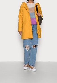 Żółta wodoodporna parka z kapturem, w zestawieniu z paskowym swetrem w pastelowych kolorach i postrzępionymi niebieskimi dżinsami. Brązowe tenisówki dopełniają stylizację.