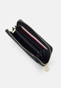 Portafoglio nero con zip e hardware dorato. Presenta diversi scomparti per carte, una tasca con zip e una texture interna liscia con accenti rossi a contrasto.