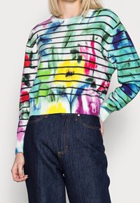 Femme portant un pull rayé coloré avec des motifs floraux abstraits et un jean bleu foncé taille haute sur un fond uni.