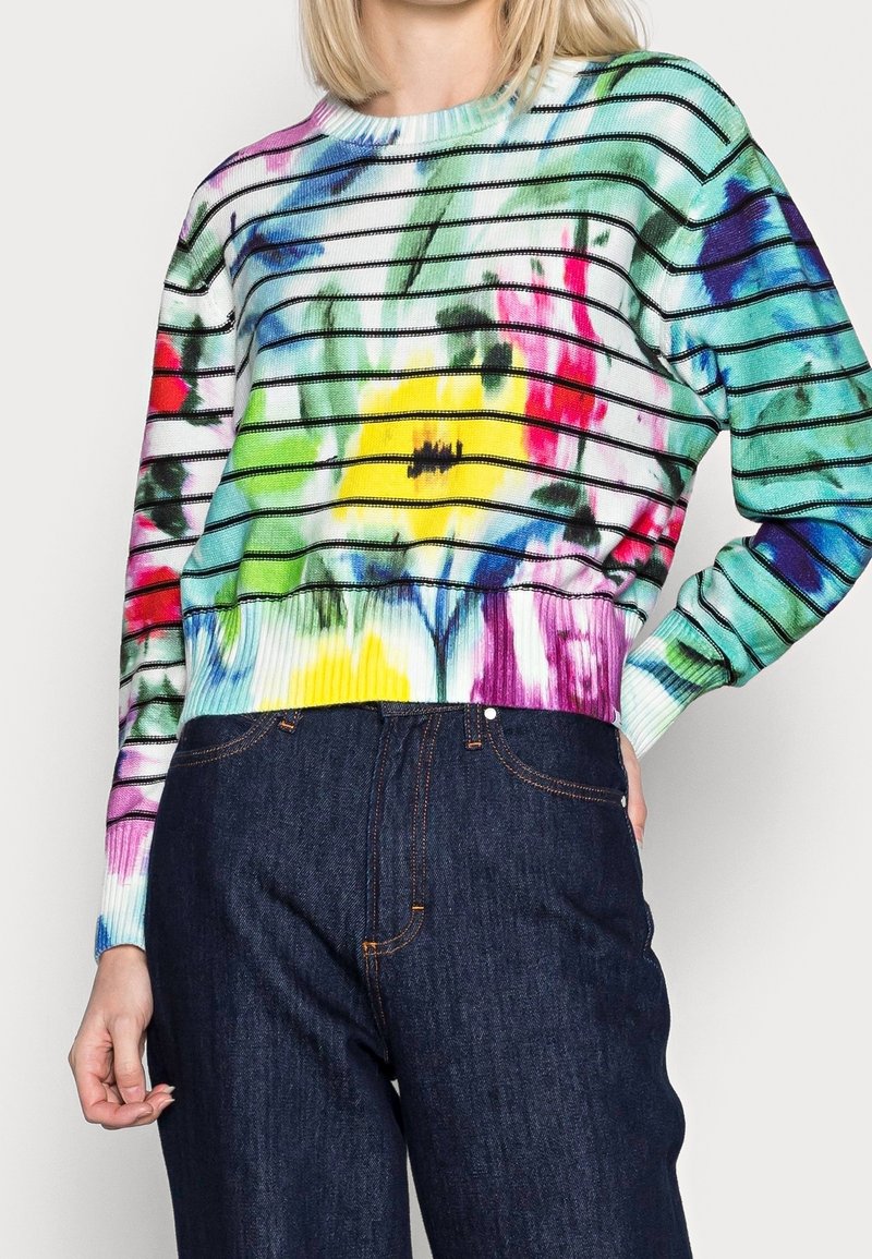 Femme portant un pull rayé coloré avec des motifs floraux abstraits et un jean bleu foncé taille haute sur un fond uni.
