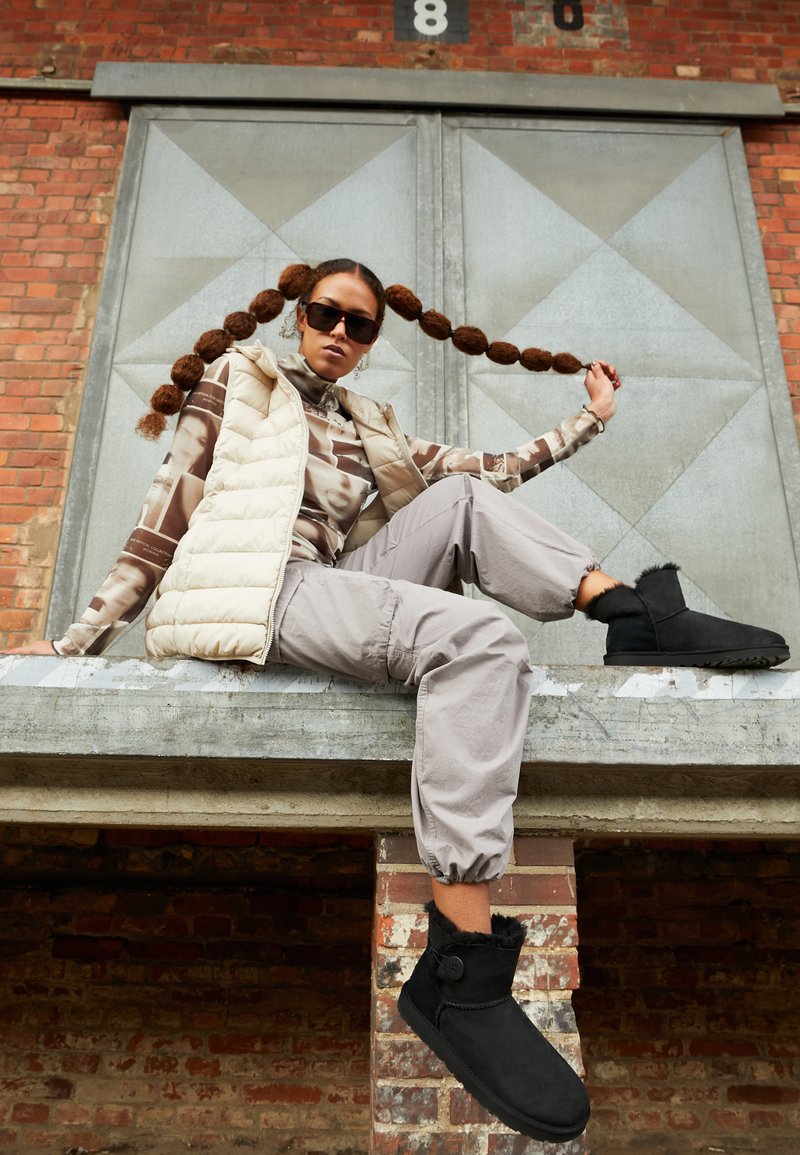 Botas negras de tobillo con acentos de pelo, chaleco acolchado beige, blusa estampada y pantalones cargo grises, contra un fondo de ladrillo texturizado y metal.