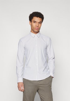 Chemise blanche à manches longues avec un motif hexagonal, col boutonné et tissu texturé. Portée avec un pantalon de couleur claire.