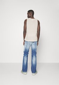 Muž otočený chrbtom, oblečený v krémovom bezrukávovom svetri, modrých vyblednutých džínoch a svetlozelených teniskách na plain bielom pozadí.