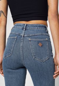 Jeans de mezclilla azul de cintura alta con un acabado texturizado, que presentan una etiqueta de cuero marrón y dos bolsillos traseros, exhibidos desde la parte posterior.