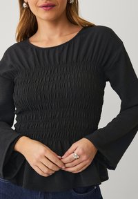 Zwarte blouse met lange mouwen en een gerimpelde bodice. Heeft een ronde halslijn en uitlopende mouwen, gemaakt van textuurstof.