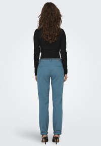 Zilas bikses ar pielāgotu piegriezumu, gludu tekstūru un šuvēm aizmugurē esošajās kabatās, apvienotas ar melnu garām piedurknēm krekla.