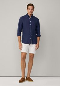 Camicia di lino blu con bottoni e maniche lunghe, abbinata a pantaloni corti bianchi e mocassini in pelle scamosciata marrone, su sfondo neutro.