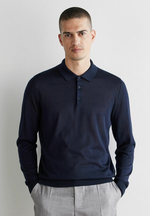 Mann mit kurzen Haaren, der einen dunkelblauen Langarm-Polo-Pullover und hellgraue Hose trägt, steht vor einer schlichten weißen Wand.