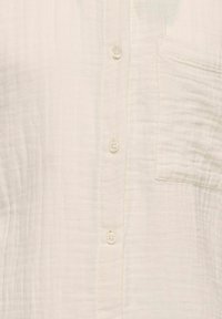 Gai krēmo pogu krekls, kas izgatavots no teksturēta auduma, ar sešām pogām un vienu krūšu kabatu.