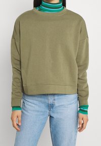 Felpa verde oliva con vestibilità ampia e scollatura a coste, sovrapposta a un dolcevita a righe teal e verde, indossata con jeans blu.