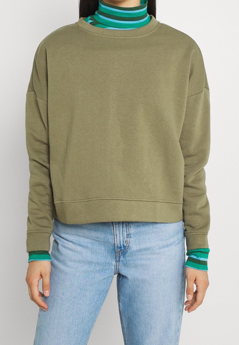 Felpa verde oliva con vestibilità ampia e scollatura a coste, sovrapposta a un dolcevita a righe teal e verde, indossata con jeans blu.