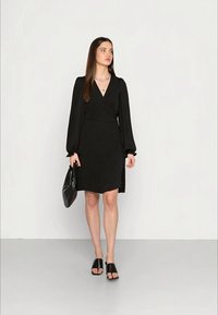 Vestido preto envolvente com mangas bufantes longas, cintura ajustada e barra drapeada. Usado com sandálias de salto baixo pretas e uma bolsa preta. Textura suave.