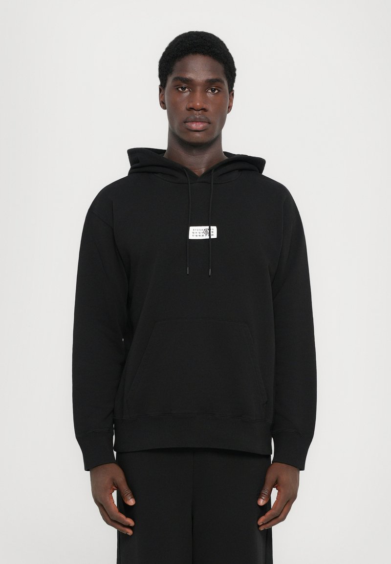 Musthaveneeru hoodie, millel on eesmine tasku, nööriga kapuuts ja väike graafiline silt rinnal. Sujuv kangas, lõdvestunud lõige ja pikad varrukad.