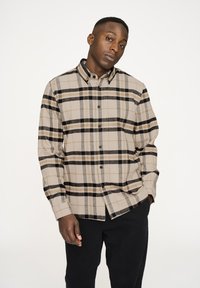 Beige und schwarze kariertes Hemd mit einem Button-Down-Kragen, langen Ärmeln und schwarzen Knöpfen, kombiniert mit dunklen Hosen. Weicher, flauschiger Stoff.