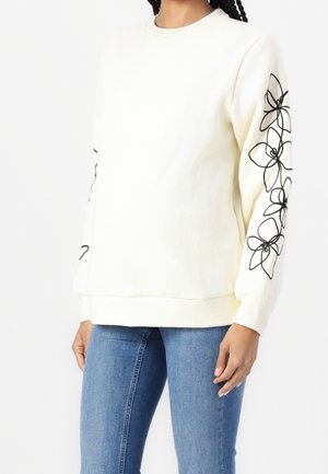 Mujer con sudadera blanca con arte lineal floral negro en las mangas y vaqueros azules, posando frente a un fondo blanco liso.