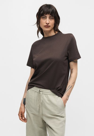 Άτομο με σκούρα μαλλιά που φορά σκούρο καφέ κοντομάνικο πουκάμισο και ανοιχτό μπεζ παντελόνι με κορδόνι, με ορατά τατουάζ στα χέρια.