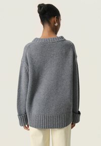 Pull gris oversize avec un col et un ourlet côtelés. Doté d'un tricot doux et texturé ainsi que d'épaules tombantes. Porté par-dessus un pantalon crème.