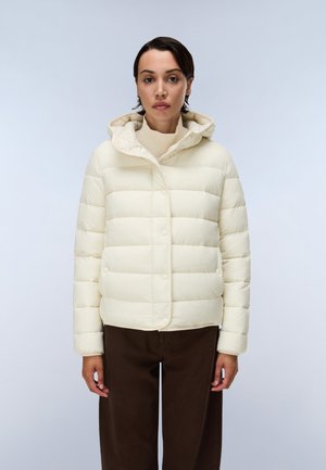Femme portant une doudoune crème avec capuche et un pantalon marron, debout devant un fond gris clair uni.