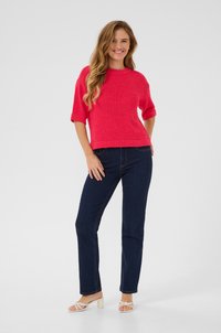 Kurzärmliger, leuchtend pinker Strickpullover, kombiniert mit dunkelblauen Jeans. Das Model trägt weiße Absatzsandalen und steht vor einem neutralen Hintergrund.
