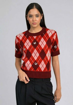 Jeune femme portant un pull à manches courtes à motif losanges rouge et rose avec des accents en bijoux, associé à un pantalon noir taille haute.