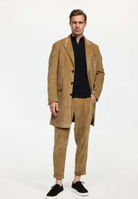 Traje de pana marrón con un abrigo largo y pantalones ajustados, combinado con una parte superior negra con cremallera y zapatillas negras. Textura suave con líneas verticales.