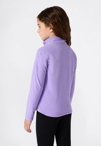 Enfant avec des cheveux bruns bouclés portant une veste en polaire lavande à manches longues et un pantalon noir, tourné de côté contre un fond blanc.