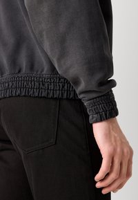 Dunkelgrauer Sweatshirt mit strukturierten, gerafften Bündchen, präsentiert neben einer schwarzen Hose. Der Stoff wirkt weich und leicht abgetragen.
