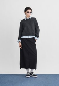 Άτομο που φοράει στρογγυλά γυαλιά ηλίου, γκρί πουλόβερ με πλέξη σε καλώδιο πάνω από μπλε πουκάμισο, μαύρη μακριά φούστα, λευκές κάλτσες και μαύρα αθλητικά παπούτσια, στέκεται σε μπλε χαλί.