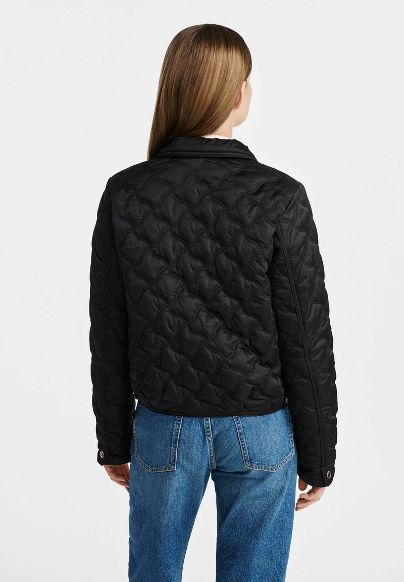 Veste matelassée noire avec un col structuré et des manches longues, arborant un motif texturé et gonflé. Associée à un jean en denim bleu clair.