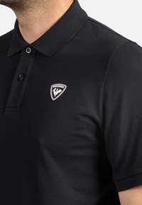 Černé polo tričko s texturovanou tkaninou, límečkem, a plaketou na tři knoflíky, s bílým logem Rossignol na levé straně hrudi.