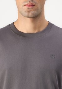 Mann trägt ein schlichtes, dunkelgraues Rundhals-T-Shirt mit kleinem, erhabenem Logo auf der linken Brust, sichtbar vom Hals bis zum oberen Torso.