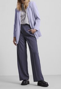 Žena v světle fialovém saku, šedém tričku, tmavě šedých širokých kalhotách a černých platformových botách, stojící s jednou rukou v kapse.