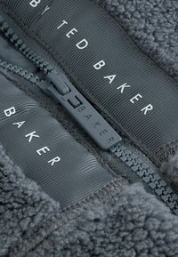 Szary polarowy materiał z prążkowymi panelami; widoczny zamek błyskawiczny z szarymi elementami; białe logo "Ted Baker" na materiale. Tekstura i miękki wygląd.