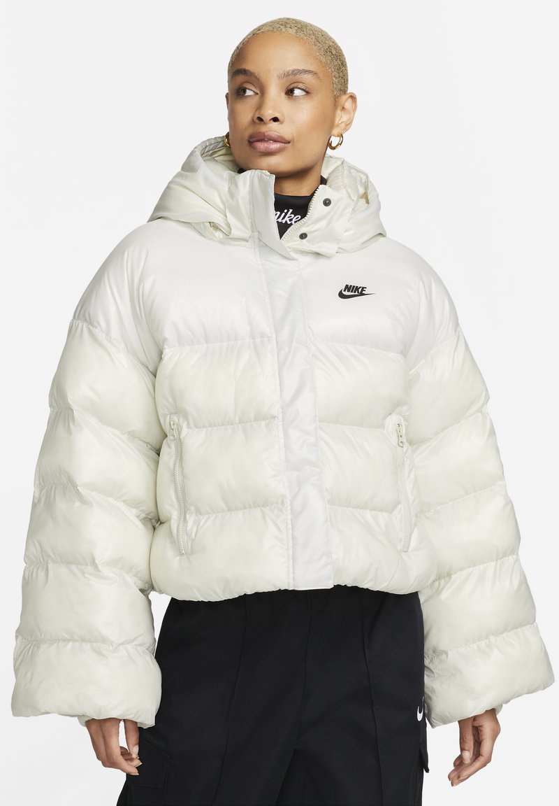 Chaqueta Nike blanca y esponjosa con capucha, que presenta costuras visibles, bolsillos con cremallera y un pequeño logo negro en el pecho.