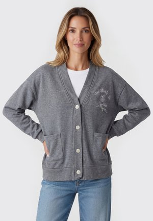 Kvinde iført grå knap-på cardigan med forlommer og broderet tekst på brystet, kombineret med lyseblå jeans.