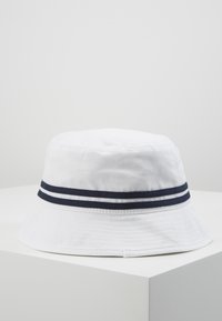 Cappello a secchiello bianco realizzato in tessuto morbido, con una striscia doppia blu navale attorno alla base. Ampia visiera inclinata e superficie liscia.