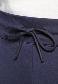 Gros plan sur la ceinture d'un pantalon de survêtement bleu marine avec un cordon noué sur un fond de chemise blanche.