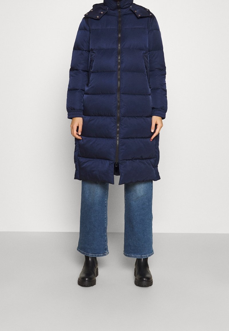 Manteau matelassé bleu marine avec fermeture zippée à l'avant, manches longues, poches latérales et détails de col boutonné, associé à un jean à jambes larges et des bottes noires.