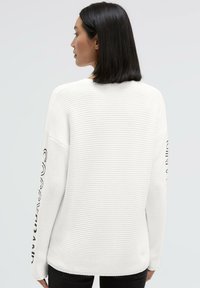 Weißer Strickpullover mit strukturiertem Muster, mit überschnittenen Schultern und schwarzer Schrift entlang des linken Ärmels. Weiches, lässiges Design.