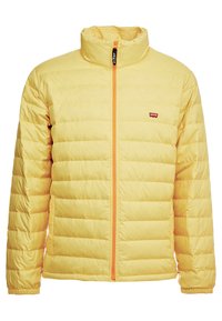Gul quiltet jakke med høj krave, lynlås foran med orange detaljer og elastiske manchetter. Lille logo på venstre bryst.