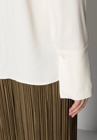 White long-sleeve blouse cuff with button over hand, paired with brown pleated skirt against neutral background.