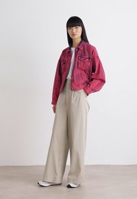 Giacca di jeans rosa con chiusura a bottoni e due tasche sul petto, abbinata a pantaloni larghi beige chiaro e sneaker bianche.