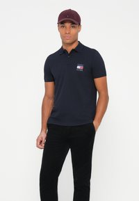 Marineblå polo skjorte med korte ermer, krage og brodert logo. I kombinasjon med svarte bukser, har modellen på seg en burgunder cap. Glatt tekstur.