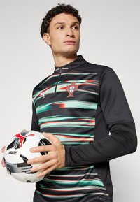 Zwarte sporttop met groene en rode golvende strepen, half-rits ontwerp en het wapen van Portugal. Model houdt een wit en zwart voetbalsysteem vast.