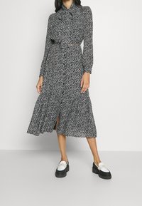 Svartvit blommig midiklänning med långa ärmar, knappfront, rynkad midja och volang i fållen; bärs med kraftiga loafers med svart sula.