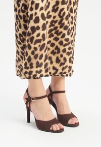 Sandalias de tacón alto de ante marrón con diseño de punta descubierta, tira en el tobillo y detalle de hebilla dorada. El fondo presenta una tela de estampado de leopardo.